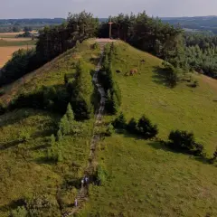 Wzgórze z drewnianym krzyżem na szczycie, schody prowadzące na górę. W oddali pola i lasy, krajobraz przypominający rezerwat przyrody cisowa.