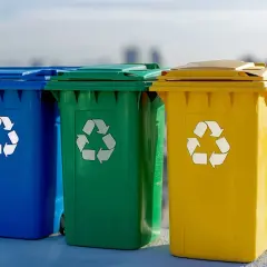 Trzy kolorowe kosze na śmieci: niebieski, zielony i żółty, z symbolami recyklingu. Uczą, jak segregować śmieci dla lepszej przyszłości.