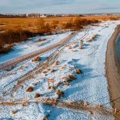 Zimowy krajobraz na rezerwacie przyrody Mechelińskie Łąki. Śnieg pokrywa plażę i wydmy, a drewniana kładka prowadzi wzdłuż morza.