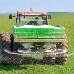 Traktor rozsypuje nawóz na zielonym polu. To przykład działań, które mogą być częścią "zielonego ładu dla rolników", mającego na celu zrównoważone rolnictwo.