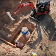 Koparka kopie dół, w którym montowana jest biała oczyszczalnia ścieków z niebieskimi rurami.