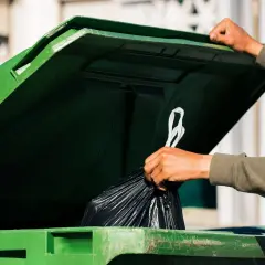 Ręka wrzuca worek ze śmieciami do zielonego kontenera. Kto jest zwolniony z opłaty za śmieci? Może ten, kto segreguje odpady.