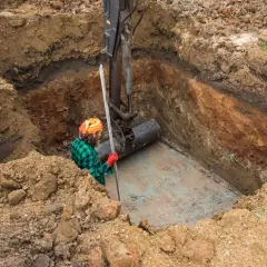 Koparka umieszcza rurę w wykopie. Pracownik w kasku i zielonej koszuli obserwuje, zastanawiając się, jaki kamień pod drenaż oczyszczalni będzie najlepszy.