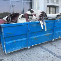 Niebieski kontener na śmieci wypełniony zrolowanymi dywanami.