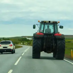 Czerwony traktor jedzie drogą, a za nim biały samochód. Widok na wiejski krajobraz. Jeśli zauważysz zanieczyszczenie drogi, wiedz, gdzie zgłosić.
