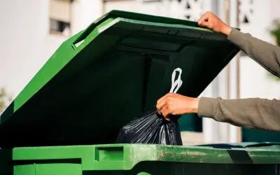 Ręka wrzuca worek ze śmieciami do zielonego kontenera. Zastanawiasz się, ile wynosi opłata za śmieci?