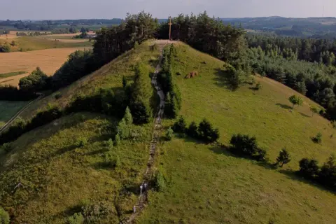 Wzgórze z drewnianym krzyżem na szczycie, schody prowadzące na górę. W oddali pola i lasy, krajobraz przypominający rezerwat przyrody cisowa.
