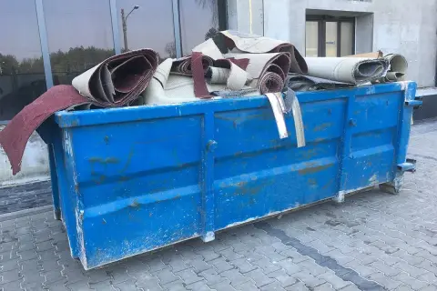 Niebieski kontener na śmieci wypełniony zrolowanymi dywanami.