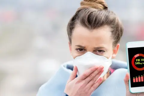 Kobieta w masce ochronnej trzyma telefon z aplikacją pokazującą wysokie stężenie pyłu PM10. Czy smog ma zapach? Ta aplikacja mówi wszystko.