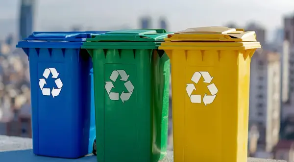 Trzy kolorowe kosze na śmieci: niebieski, zielony i żółty, z symbolami recyklingu. Uczą, jak segregować śmieci dla lepszej przyszłości.