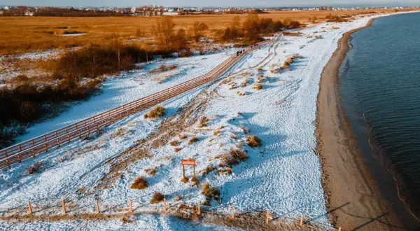 Zimowy krajobraz na rezerwacie przyrody Mechelińskie Łąki. Śnieg pokrywa plażę i wydmy, a drewniana kładka prowadzi wzdłuż morza.