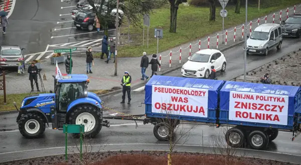 Niebieski traktor ciągnie przyczepę z banerami "Pseudoekolodzy was oszukują" i "Unijna polityka zniszczy rolnika".