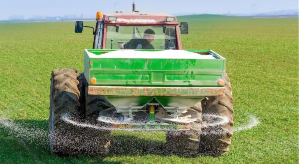 Traktor rozsypuje nawóz na zielonym polu. To przykład działań, które mogą być częścią "zielonego ładu dla rolników", mającego na celu zrównoważone rolnictwo.