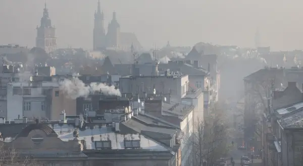 Widok na miasto w smogu z dymiącymi kominami i zarysami wież kościelnych w tle.
