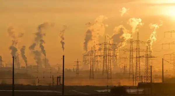 Dymiące kominy fabryk i linie energetyczne na tle pomarańczowego, zachodzącego słońca.