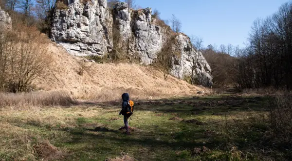 Dziecko z plecakiem przemierza rezerwat przyrody Dolina Kluczwody, podziwiając skalne ściany i wiosenną przyrodę.