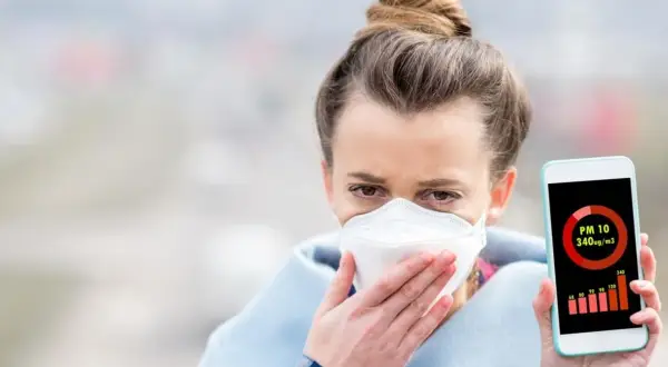 Kobieta w masce ochronnej trzyma telefon z aplikacją pokazującą wysokie stężenie pyłu PM10. Czy smog ma zapach? Ta aplikacja mówi wszystko.