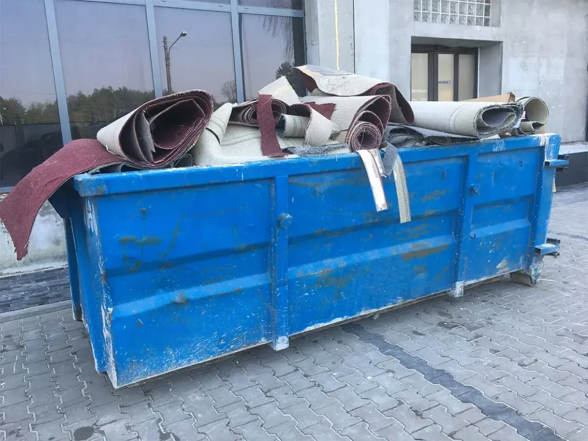 Niebieski kontener na śmieci wypełniony zrolowanymi dywanami.