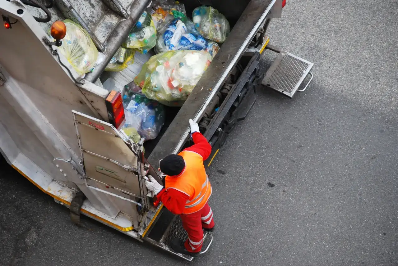 Śmieciarka z workami pełnymi odpadów. Pracownik w kamizelce odblaskowej i czerwonym stroju przygotowuje się do załadunku. Zastanawiasz się, ile kosztuje tona śmieci na wysypisku?