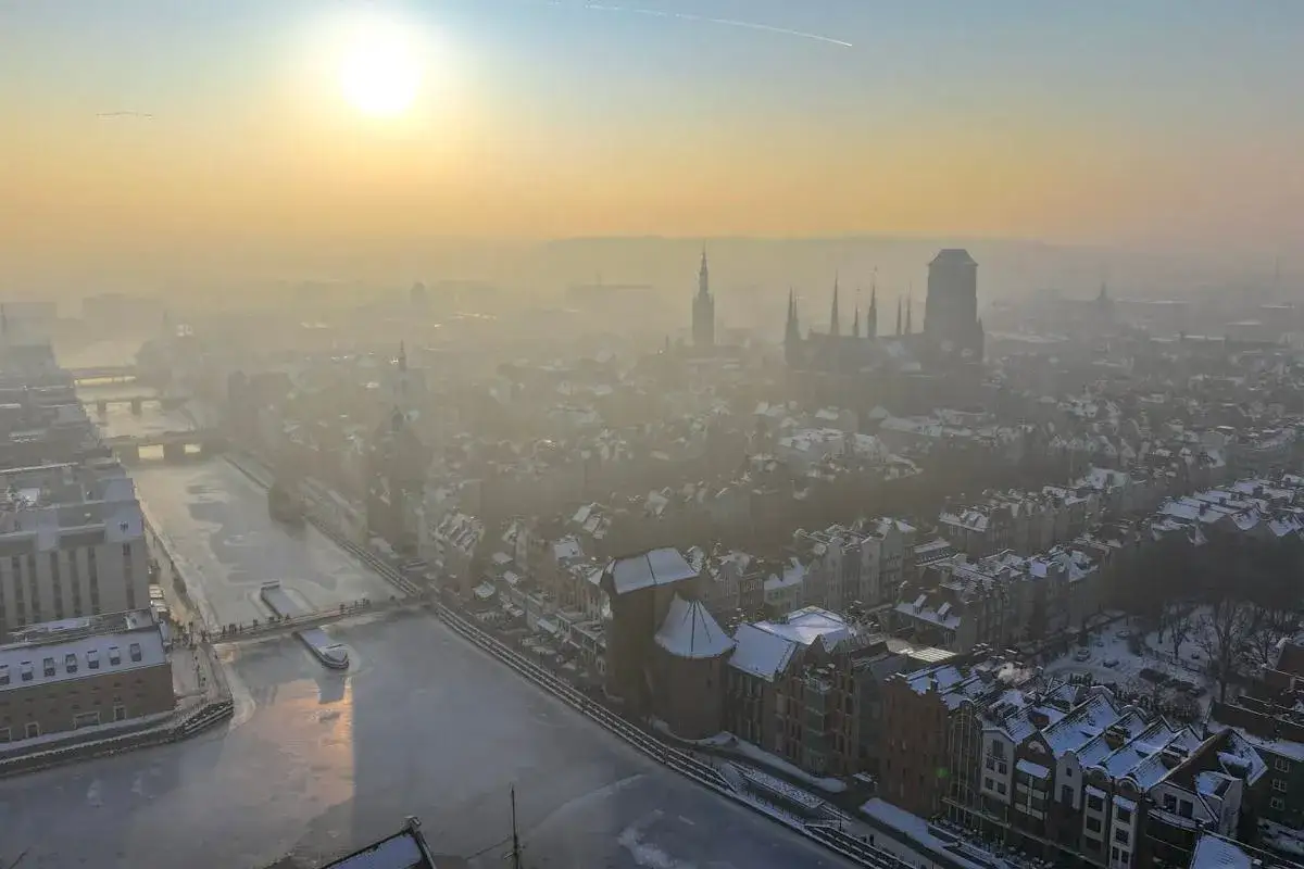 Zimowy dzień w mieście, słońce ledwo przebija się przez gęsty, żółtawy welon. Widać zamarzniętą rzekę, zabudowania i wieże kościołów. Obraz pokazuje, jak powstaje smog.