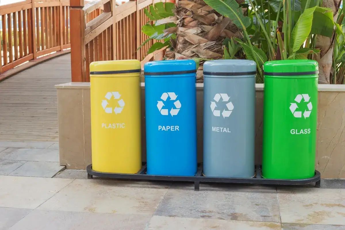 Cztery kolorowe kosze (żółty, niebieski, szary, zielony) z symbolami recyklingu i napisami: PLASTIC, PAPER, METAL, GLASS. Uczą, jak segregować śmieci.
