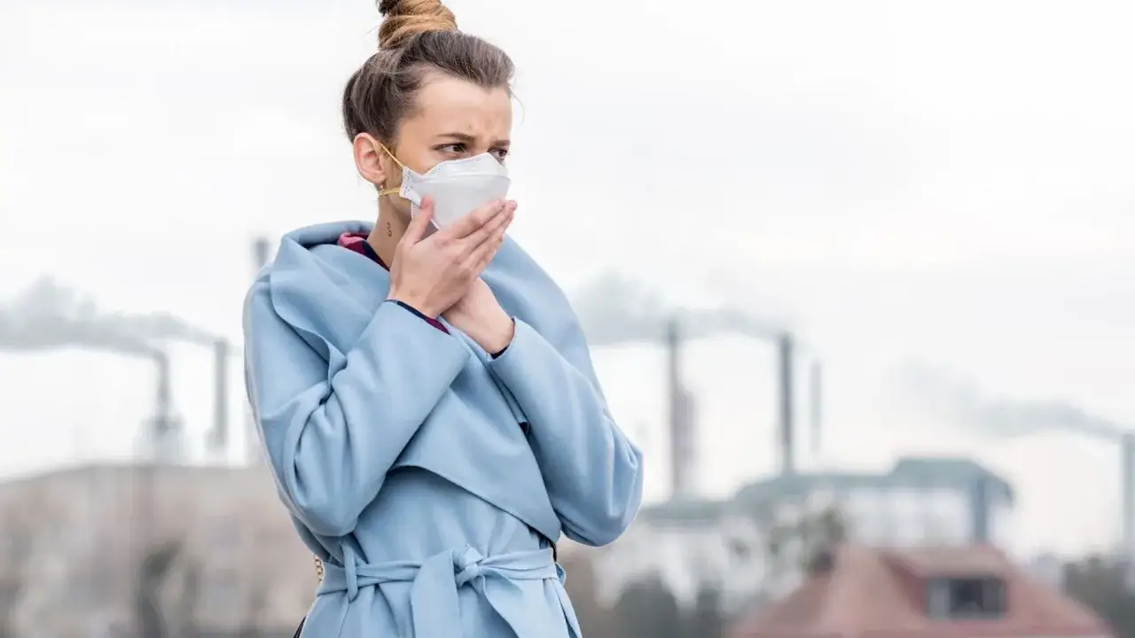 Kobieta w masce ochronnej kaszle na tle dymiących kominów. Jak leczyć zatrucie smogiem? Unikaj zanieczyszczonego powietrza.