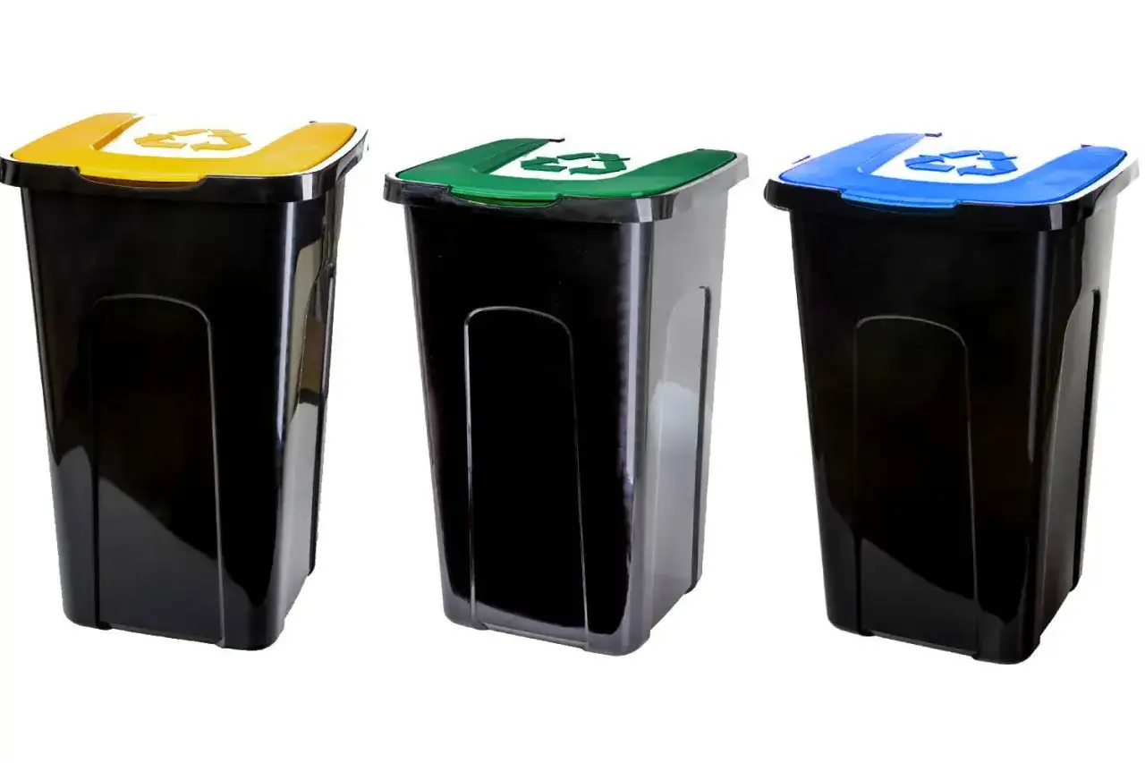 Trzy czarne kosze na śmieci z kolorowymi pokrywami: żółtą, zieloną i niebieską. Każda pokrywa ma symbol recyklingu. To niebieski pojemnik na śmieci, do którego wrzucamy papier.