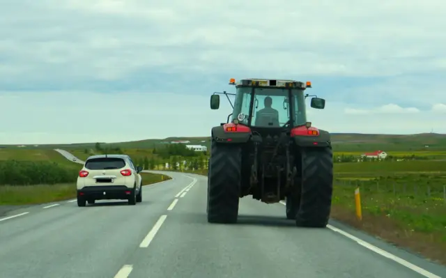 Czerwony traktor jedzie drogą, a za nim biały samochód. Widok na wiejski krajobraz. Jeśli zauważysz zanieczyszczenie drogi, wiedz, gdzie zgłosić.